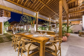 An outdoor dining area with wooden tables and chairs, surrounded by plants and a pool. - Villa Hoi in Seminyak (Seminyak)