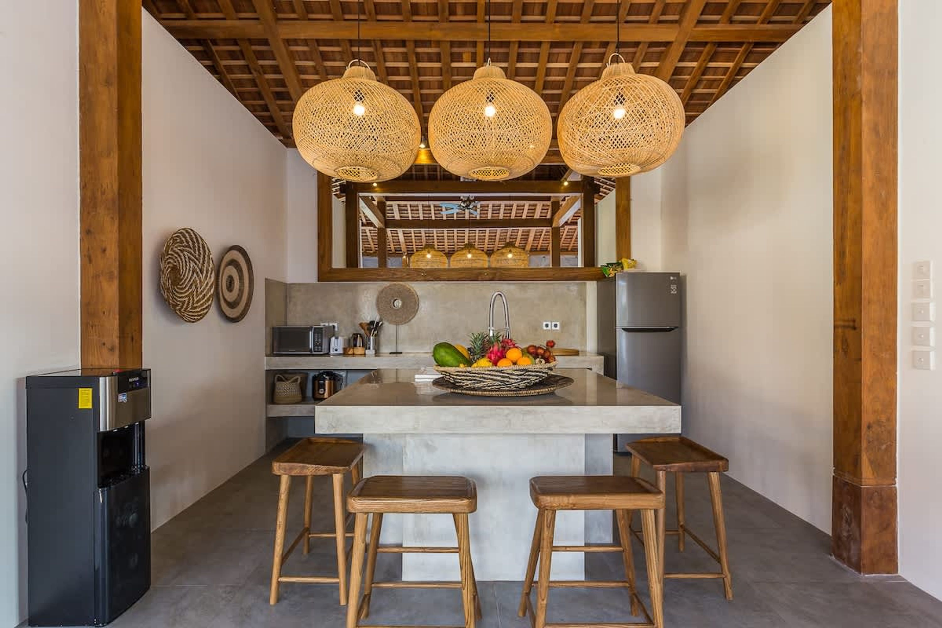 Une cuisine dotée d’un îlot central, de tabourets en bois, d’un réfrigérateur et de luminaires suspendus en osier.