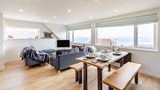Un salon moderne avec un canapé, une table à manger pour deux personnes, un téléviseur à écran plat et de grandes fenêtres offrant une vue sur la mer.