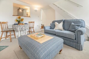 Ein modernes Wohnzimmer mit einem blauen Sofa, Holzstühlen, einem Esstisch mit floralem Tischläufer und einer Treppe.