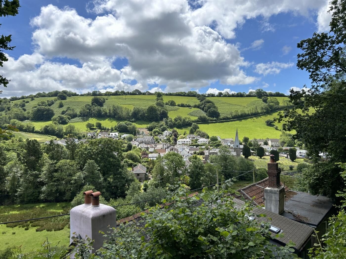 A rural village with houses, a church, and rolling green hills under a partly cloudy sky.