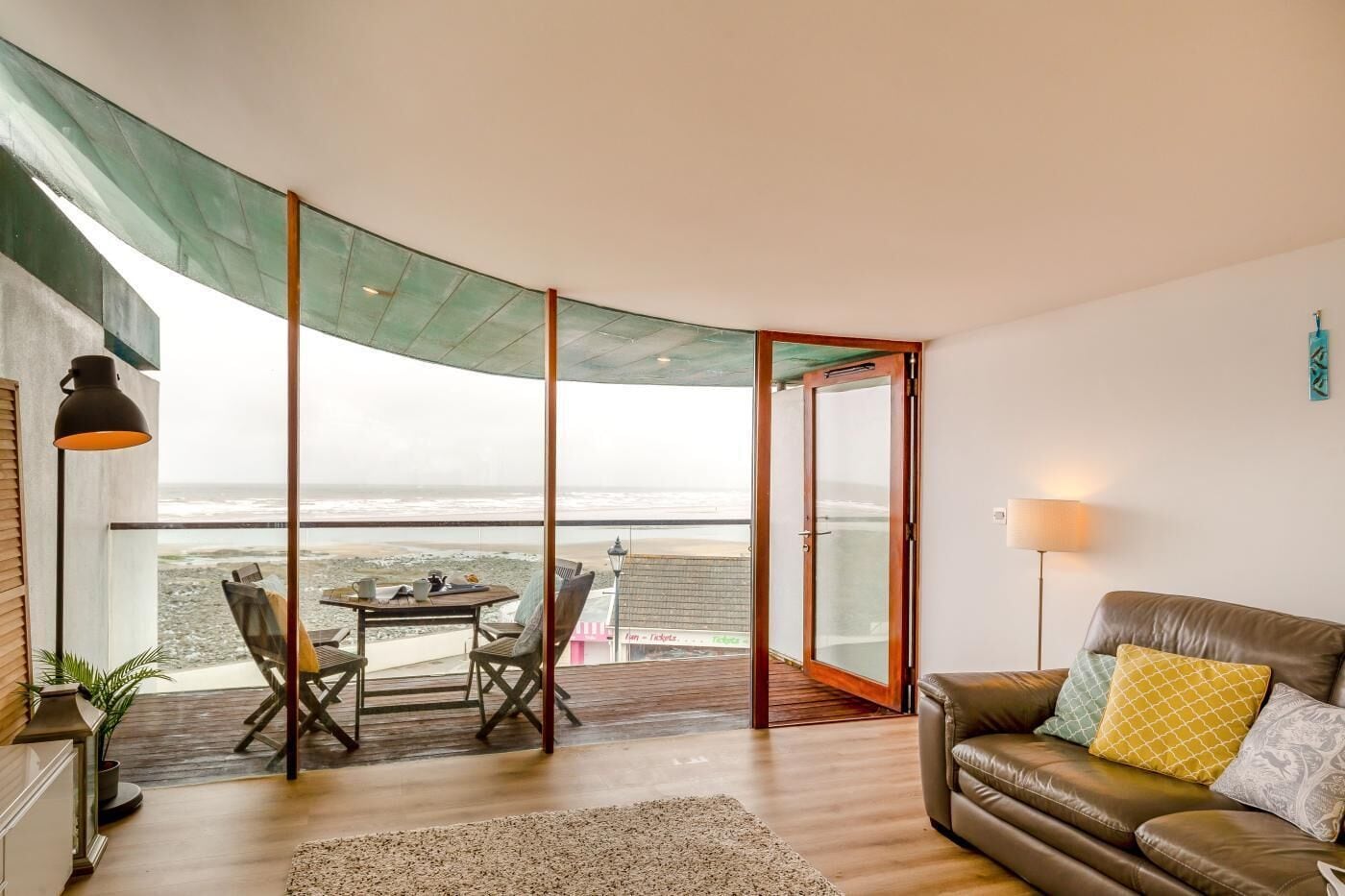 Une pièce dotée d’un plafond incurvé, d’une porte vitrée donnant sur un balcon avec vue sur la mer, d’un canapé en cuir agrémenté de coussins et d’une table avec des chaises.