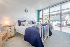 A bedroom with a large window, a bed with a white headboard, a bedside table with a lamp, and a view of a body of water and buildings. - 2 Bed in Westward Ho (oc-n44873) (Westward Ho!)