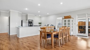 A modern kitchen with white cabinetry, a dining area with wooden chairs and a table, and a view of a backyard through a glass door.