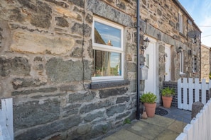 Ein Steinhaus mit einem weißen Fenster, einer Tür und einem kleinen Vorbereich mit Topfpflanzen.