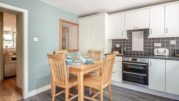 Cocina moderna con armarios blancos, salpicadero de azulejos negros y un comedor con mesa y sillas de madera.