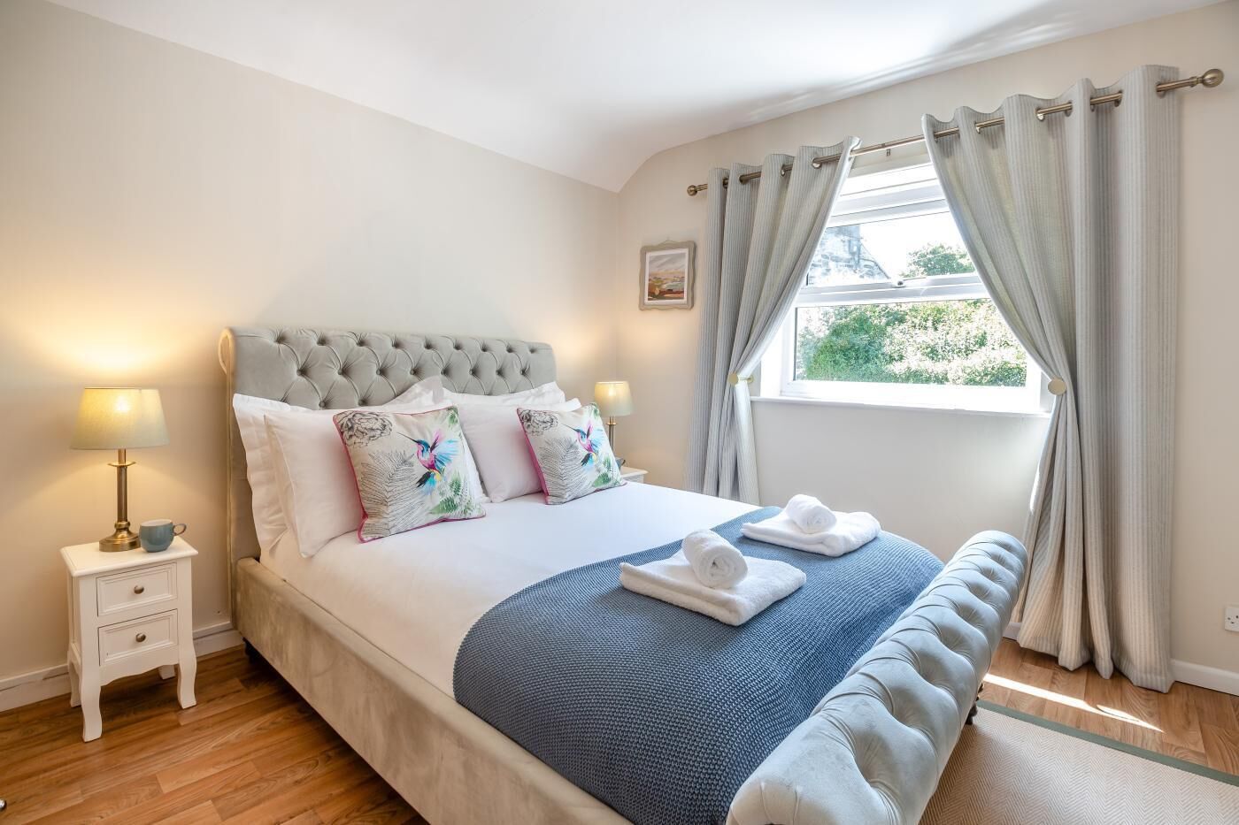 A neatly arranged bedroom with a tufted headboard, a bed with white linens, decorative pillows, a bedside table with a lamp, and a window with curtains.