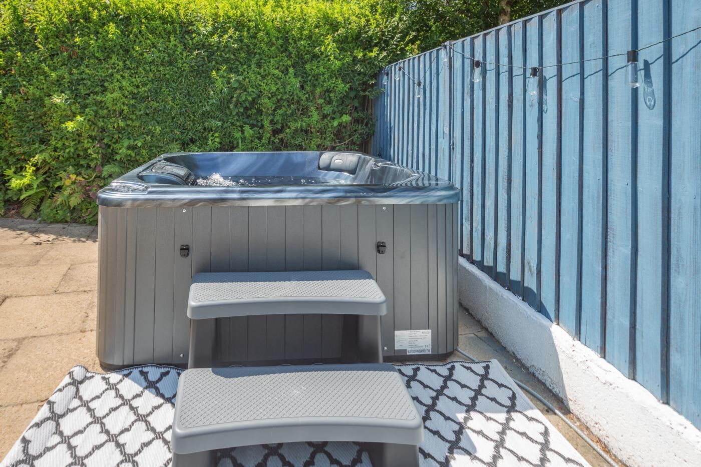 A hot tub with a step and a bench, surrounded by a blue fence and greenery.