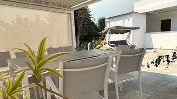 Eine Terrasse im Freien mit Tisch und Stühlen, einem überdachten Bereich und Blick auf ein Gebäude im Hintergrund.