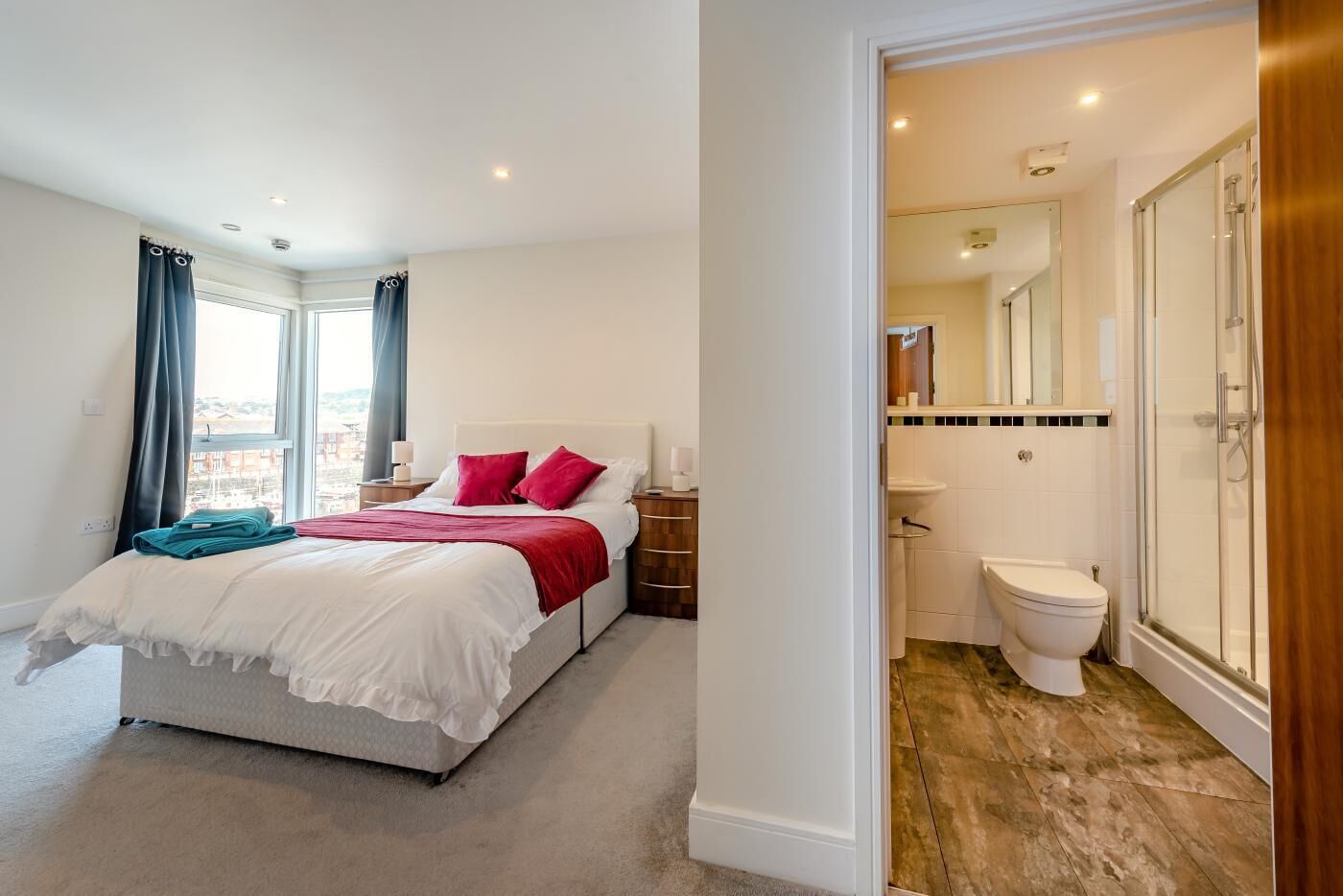 A hotel room with a bed, a window with curtains, a bathroom with a toilet and shower, and a view of the city.