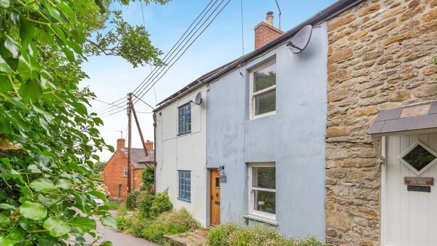 A narrow street with traditional stone and painted buildings, a visible satellite dish, and a small garden area with flowers. - 2 Bed in Eype (oc-l42804) (Eype)