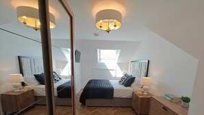A modern bedroom with a herringbone floor, a large bed with a dark headboard, bedside tables with lamps, and a large mirror. - 4 Bed in Hampeth (oc-w45221) (Shilbottle)