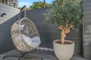Una silla colgante con un cojín, un árbol en maceta y una pared gris.