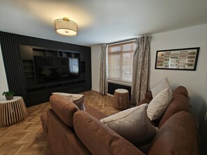 Een moderne woonkamer met een flatscreen televisie, houten vloeren, een zithoek en een behang met patroon.