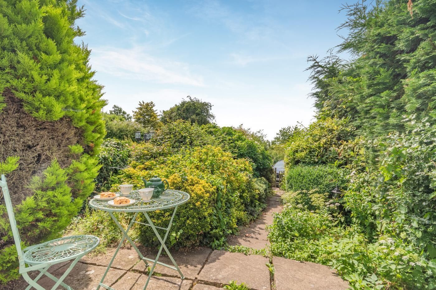 A garden pathway with a metal table and chairs, surrounded by dense foliage and a greenhouse in the distance.