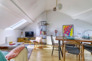 Ein Loft mit schrägem Dach, Essbereich, Fernseher, Bücherregal und einem farbenfrohen abstrakten Gemälde.