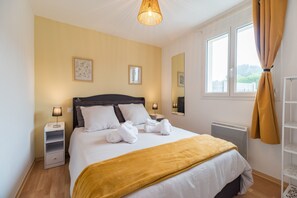 Ett hotellrum med en säng, sänglampor, ett fönster med gardiner, ett element och en hylla med handdukar.