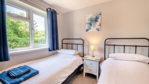 A bedroom with two beds, a window with curtains, a nightstand with a lamp, and a painting on the wall. - 2 Bed in Brixham (oc-k45223) (Brixham)