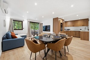 Ein modernes Wohnzimmer mit Sofa, Esstisch und Stühlen. Es gibt eine Küchenzeile mit Holzschränken und einem Flachbildfernseher.