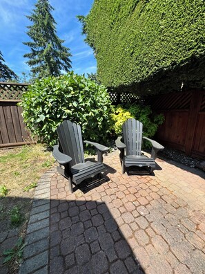 Dos sillas Adirondack en un patio empedrado con una cerca de madera y un arbusto bien cuidado al fondo.