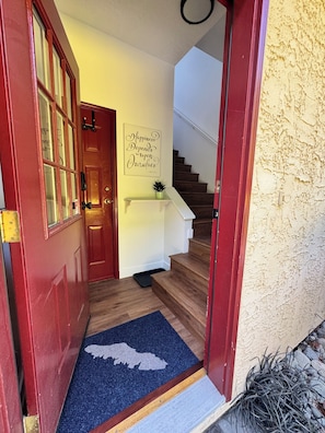 Una puerta roja con paneles de vidrio, un tapete azul con silueta blanca, un banco blanco, una escalera y una pared con una cita.