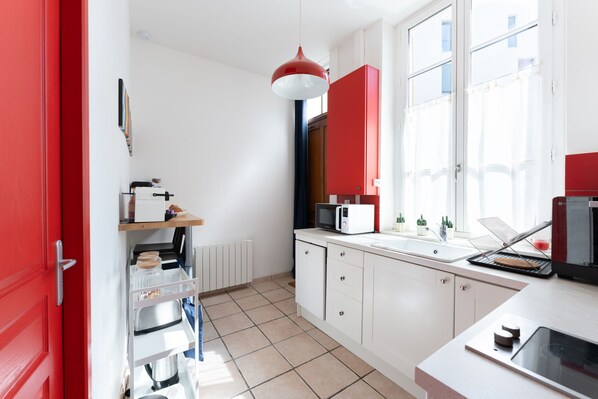 A kitchen with white cabinets, a red door, a microwave, and a window with a view. - Le Verdun - Appartement cosy proche Croix-Rousse (Caluire-et-Cuire)