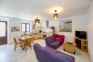 A modern living room with a purple sofa, wooden dining table, and a kitchen in the background. - L’Escapade Lanvollonnaise - Plage à 15 min (Lanvollon)