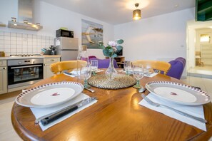 A dining table set with plates, cutlery, and glasses, surrounded by chairs, in a modern kitchen with appliances and a painting on the wall. - L’Escapade Lanvollonnaise - Plage à 15 min (Lanvollon)