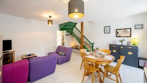 A modern living room with a purple sofa, a dining table with chairs, a staircase with green railings, and a TV on the left. - L’Escapade Lanvollonnaise - Plage à 15 min (Lanvollon)