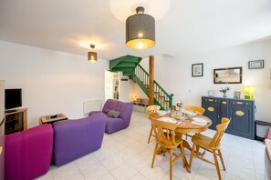 A modern living room with a purple sofa, a dining table with chairs, a staircase with green railings, and a TV on the left. - L’Escapade Lanvollonnaise - Plage à 15 min (Lanvollon)