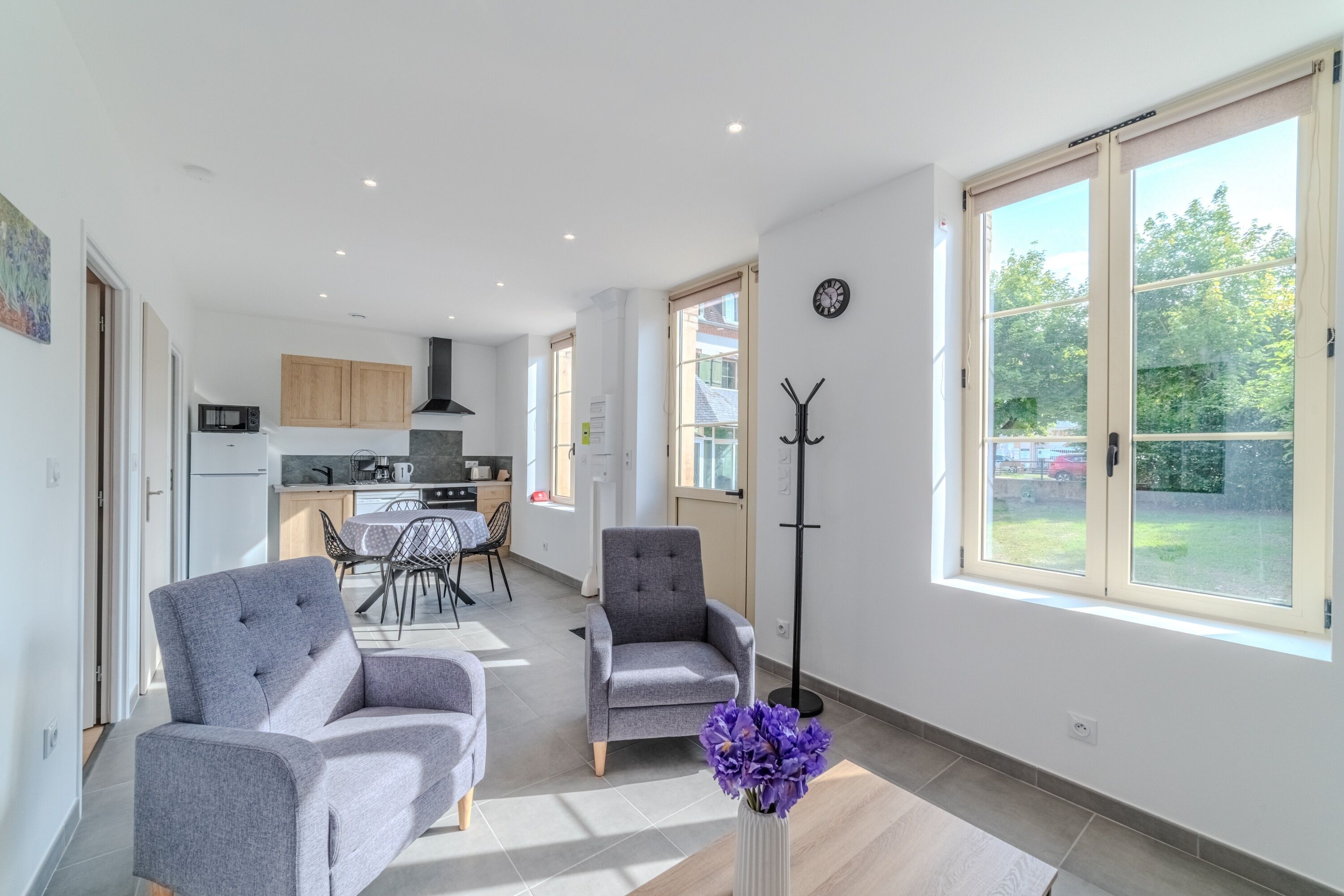 A spacious living area with a sofa, armchair, and a floor lamp. There is a dining table with chairs, a kitchen area, and large windows with a view of the outdoors.