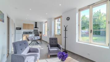 A spacious living area with a sofa, armchair, and a floor lamp. There is a dining table with chairs, a kitchen area, and large windows with a view of the outdoors.