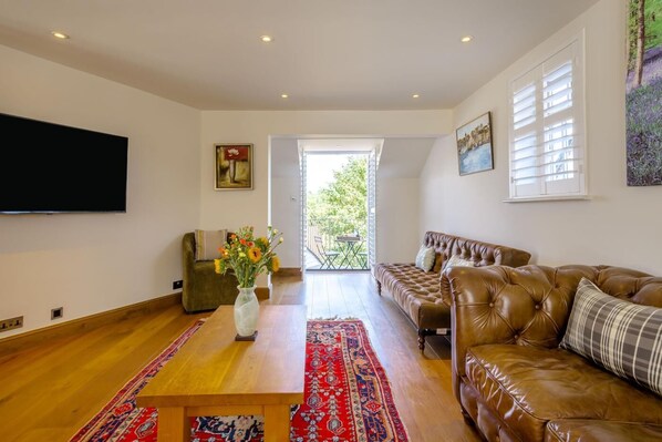 Un salón amplio con un televisor de pantalla plana grande, un sofá de cuero marrón, un sillón verde y una mesa de centro de madera con un jarrón de flores.