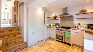 Een moderne keuken met een gasfornuis, oven en een inbouwafzuigkap. Er zijn witte kasten en een houten trap die naar een andere kamer leidt.