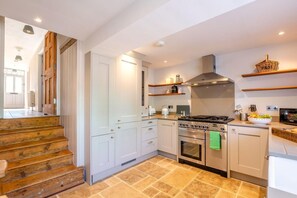 A modern kitchen with a gas stove, oven, and a built-in range hood. There are white cabinets and a wooden staircase leading to another room. - 3 Bed in Forest Row (oc-h43282) (Forest Row)