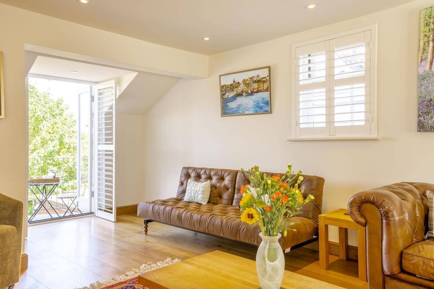 A spacious living room with a brown leather sofa, a wooden coffee table, and a vase of flowers. There is a painting of a coastal scene on the wall, and a window with blinds.