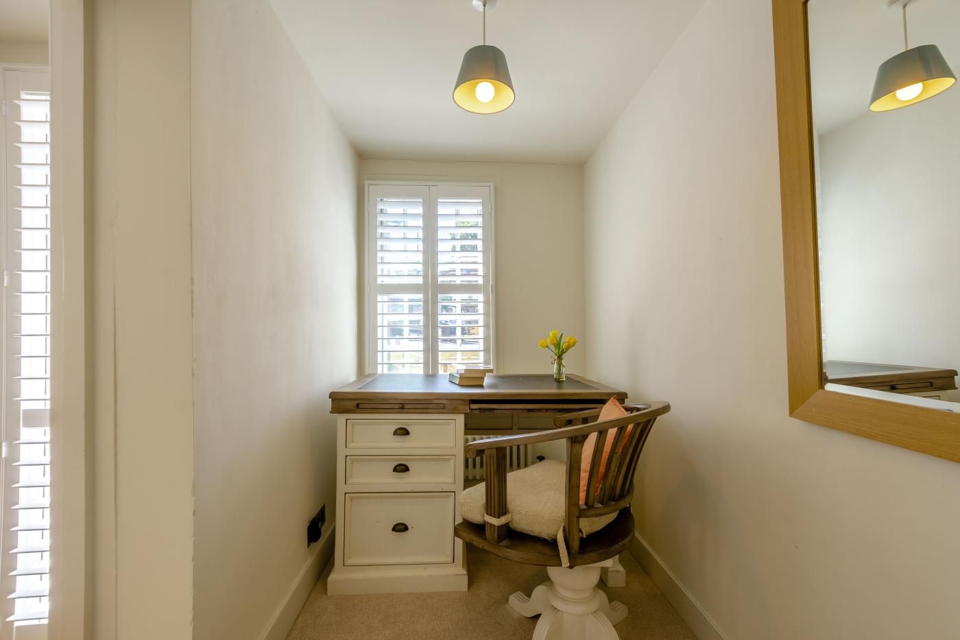 A narrow hallway with a desk and chair, a window with blinds, and a mirror on the wall.