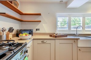 Una cocina con estufa a gas, microondas, un tazón de fruta y una ventana con persianas.
