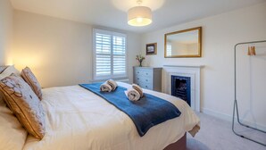 Een slaapkamer met een bed, een dressoir, een spiegel, een open haard en een raam met jaloezieën.