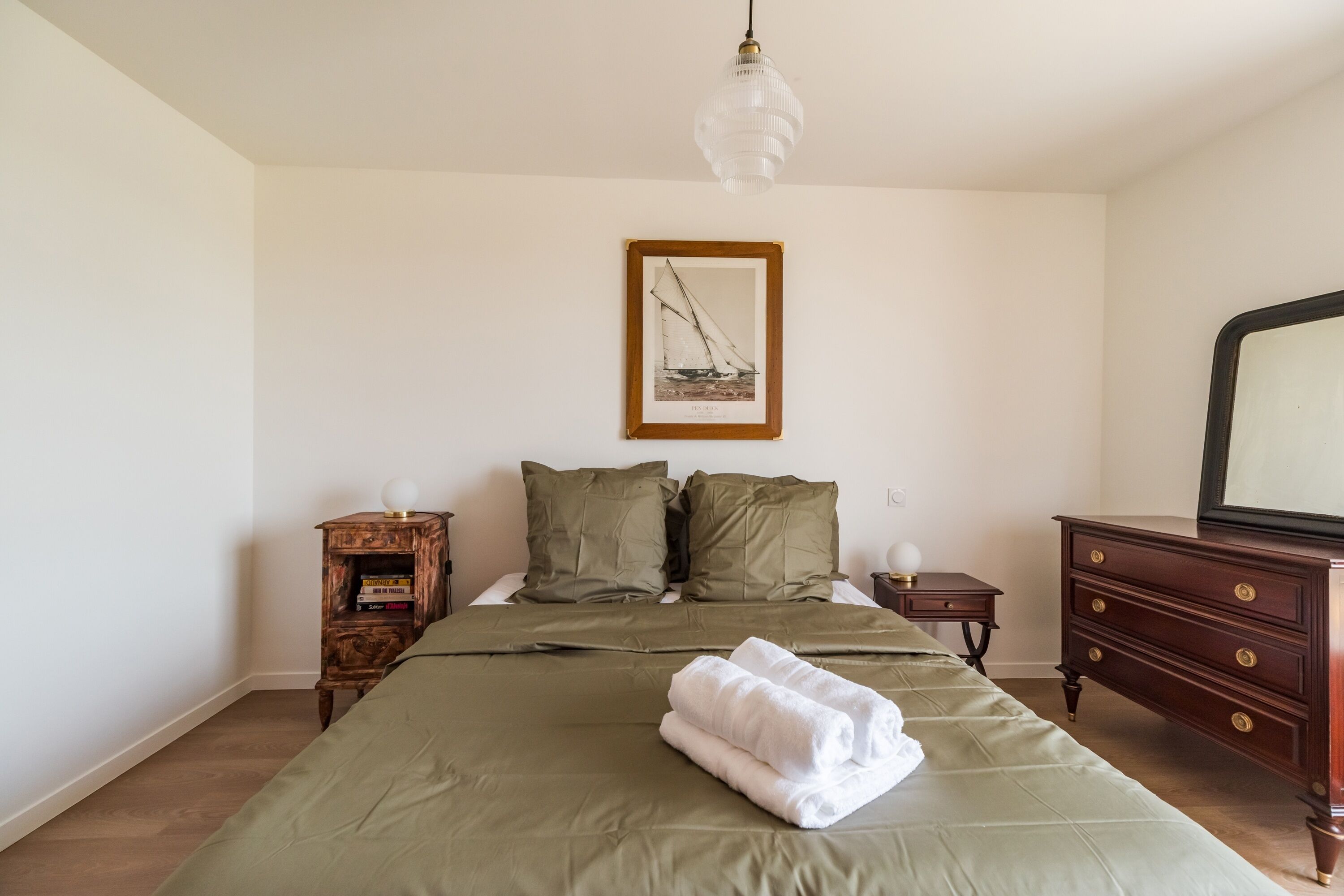 Une chambre à coucher avec un lit, des oreillers, une commode, une table de chevet, un tableau encadré au mur et un luminaire suspendu.