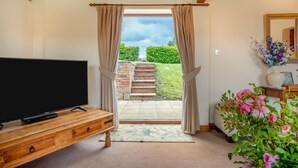 A room with a TV on a wooden cabinet, a door leading outside, a mirror, and a vase with flowers. - 2 Bed in Stratford -upon- Avon (oc-cc009) (Welford-on-Avon)
