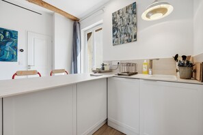 Eine moderne Küche mit weißen Schränken, einem Holzbalken an der Decke, einer Hängeleuchte und einer Tür mit einem blauen, abstrakten Gemälde.