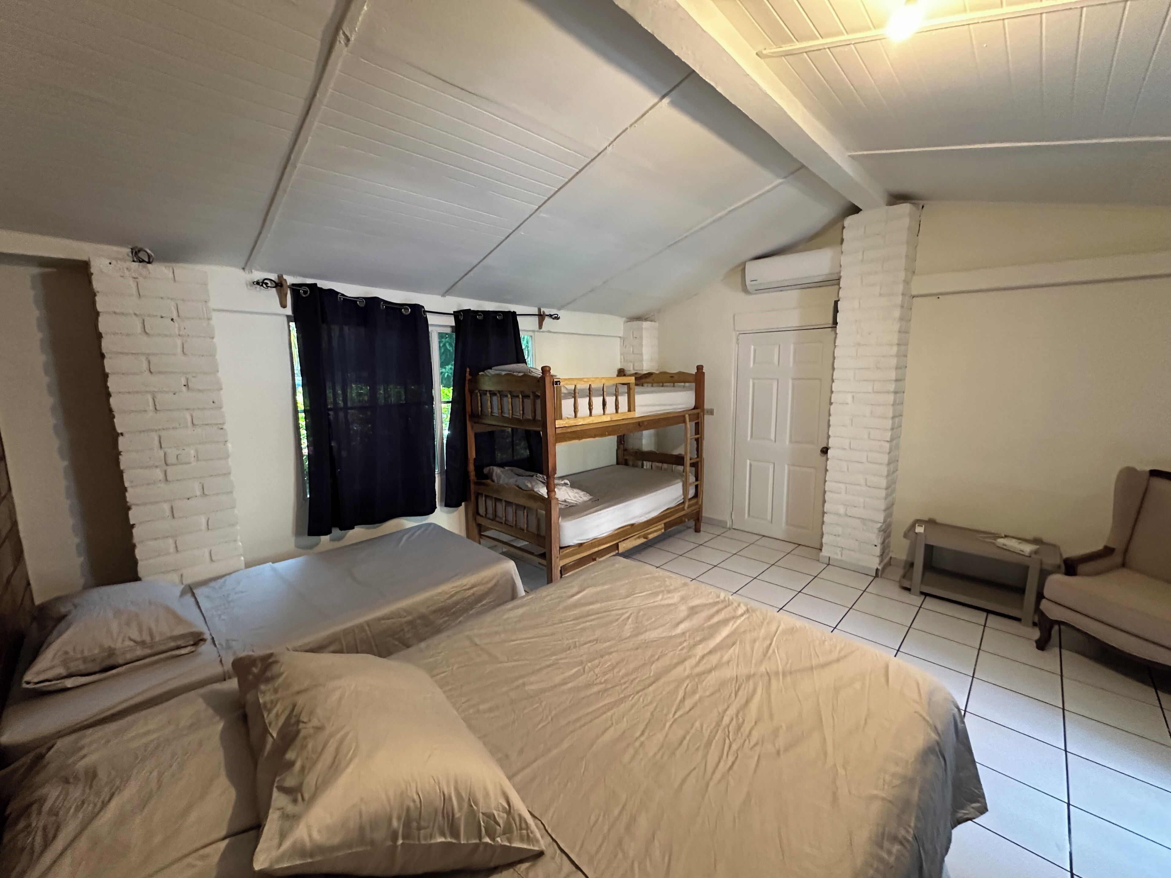 Quarto com duas camas, uma cama beliche, uma cadeira e uma porta.