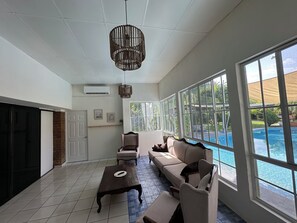 A spacious living room with a large window overlooking a swimming pool, a fireplace, and a ceiling fan. - Palm beach  (San Diego)