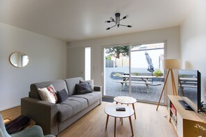 Ein modernes Wohnzimmer mit einer Couch, zwei Couchtischen, einem Fernseher und einer großen Schiebeglastür, die zu einem Außenbereich führt.