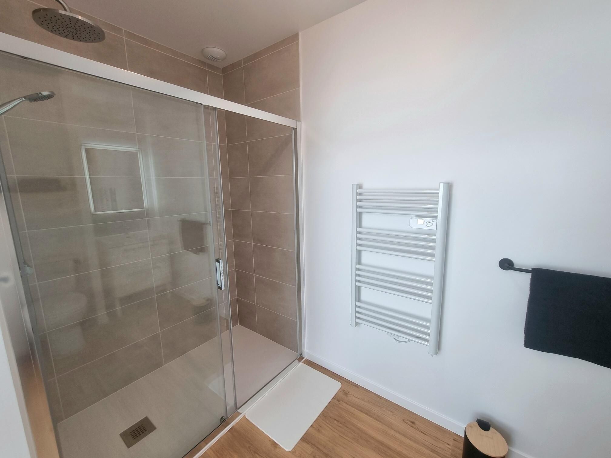 Un baño moderno con cabina de ducha de vidrio, un perchero blanco y suelo de madera.