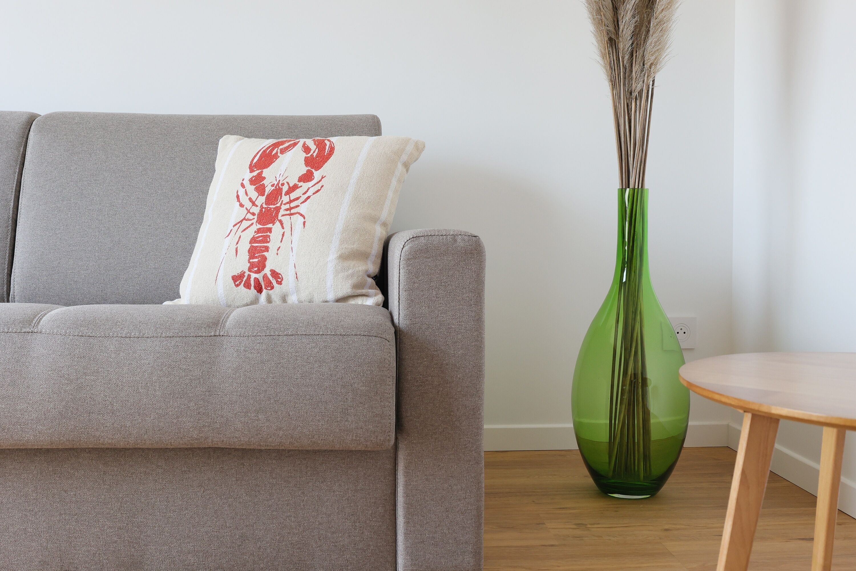 A grey sofa with a red lobster pillow, a green vase with pampas grass, and a wooden table.