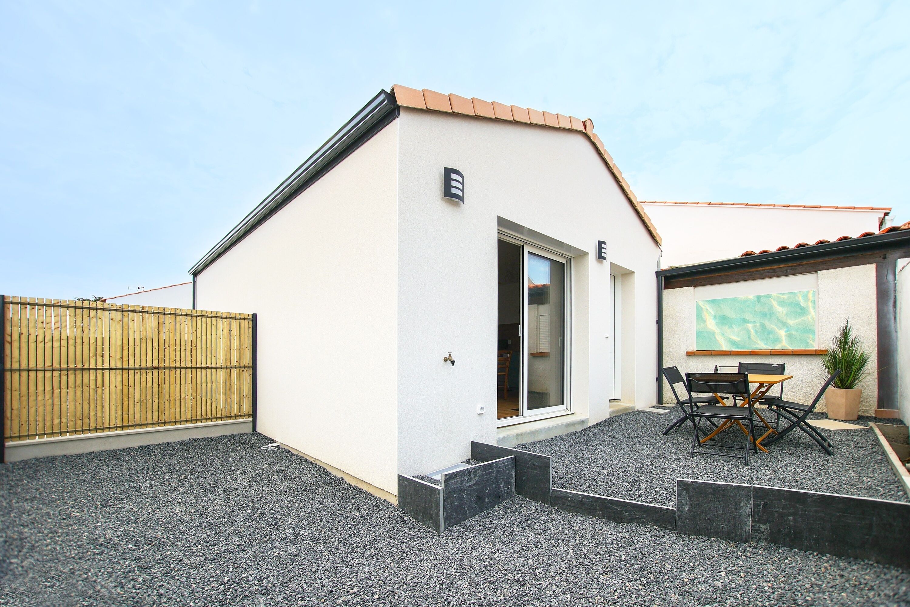 Ein modernes Gebäude mit weißer Fassade, einem Holzzaun, einer gepflegten Schotterterrasse und einem Tisch mit Stühlen.