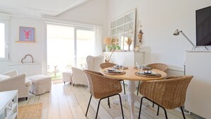 Uma sala de estar e jantar ampla e luminosa, com móveis de vime, uma grande janela e uma televisão de tela plana fixada na parede.