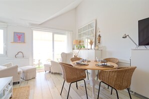 A bright, open-plan living and dining area with wicker furniture, a large window, and a flat-screen TV mounted on the wall. - Le 21 des dunes - La mer comme horizon (Saint-Jean-de-la-Rivière)